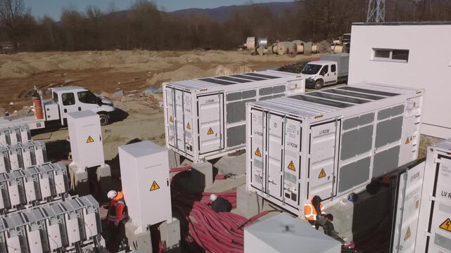 Drone aerial close view of containerized battery energy storage system (BESS) under construction with inverter units, battery racks and heavy DC cable runs at grid site.