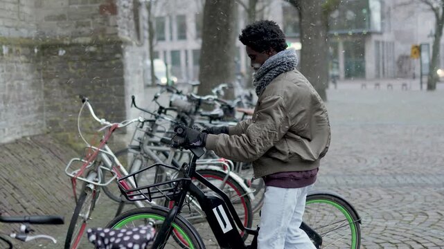 Black man unlocking a shared e-bike from a parking station in a european city during a winter snow flurry