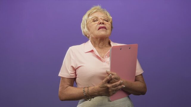 Woman holding pink clipboard with clasped hands and head tilted upward in a studio; confidence dignity poise.