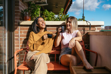 Lesbian couple laughing enjoying morning coffee on balcony