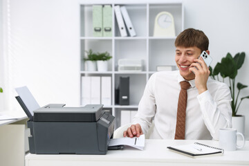 Fototapeta premium Smiling man taking printed sheet of paper from printer and talking by smartphone at white desk in office