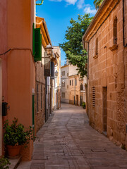 Fototapeta premium Narrow cobblestone alley winds through traditional Mediterranean buildings in Alcudia's old town, showcasing classic Mallorcan sandstone construction and peaceful residential charm.