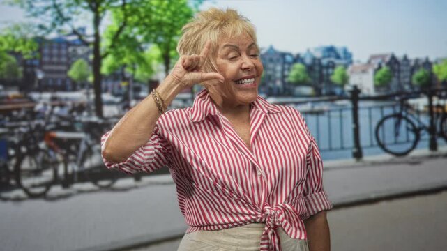 Woman forms l shape with hand by temple on street beside canal railing and parked bicycles, smiling with closed eyes in sunny urban scene; joy playfulness.