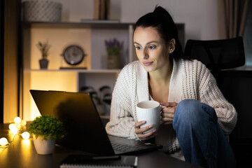 A woman enjoying a relaxed evening with a laptop while sipping from a warm cup in dim lighting. © Dejan