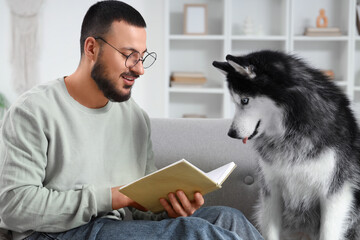 Handsome young man lying on sofa and reading book with cute husky dog at home © Pixel-Shot