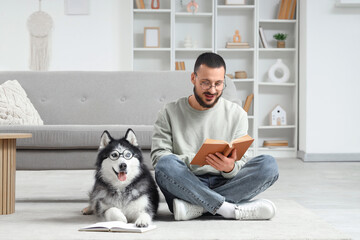 Handsome young man lying on floor and reading book with funny husky dog in eyeglasses at home © Pixel-Shot