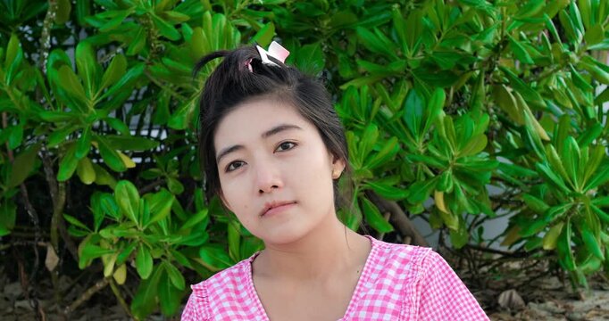 Young asian woman smiling outdoors by lush foliage wearing pink gingham dress and hair clip, candid headandshoulders portrait against tropical shrub backdrop, soft natural light, relaxed friendly