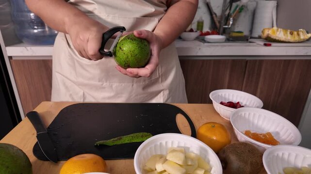 A woman in a apron prepares a fruit salad, peels and slices the avocado into cubes with a knife and pours it into a bowl.