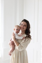 Smiling mother in dress holding her cute baby near white wall at home
