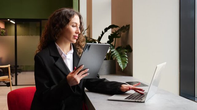 Woman uses tablet while seated at desk with laptop during work hours in office area