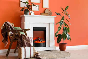 Fototapeta premium Fireplace with armchair, houseplants and photo frames near orange wall in living room