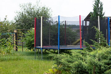Fototapeta premium Backyard playground with trampoline and protective safety net in green garden near family house on sunny summer day. Safe active games, child protection, outdoor fitness, healthy lifestyle and fun