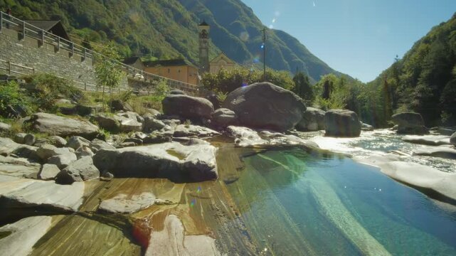 Smooth cinematic lifting shot revealing a beautiful river with crystal clear waters in Lavertezzo, Switzerland with an old town and mountains in the background.