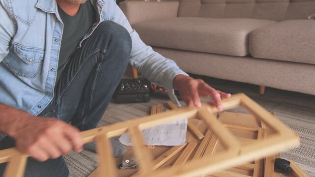 Focused man carefully reading instructions and assembling new wooden furniture in his living room