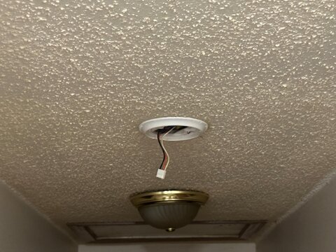 Close-up of an open recessed ceiling light fixture showing exposed electrical wiring and mounting hardware against a textured popcorn ceiling. Home maintenance, repair, and electrical installation