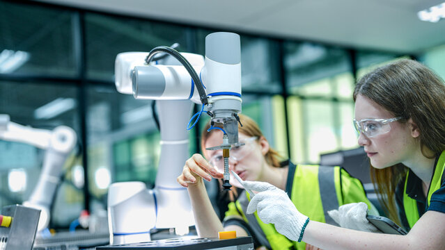 Two female engineers in safety vests collaborate in a tech lab. One woman in gloves inspects a high tech robotic arm while her colleague takes notes.