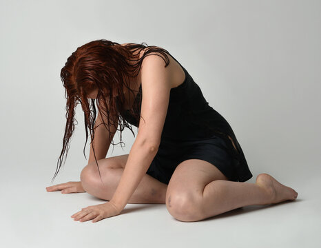 full length portrait of beautiful female model with wet messy hair and simple black silk robe gown, bruised legs. sitting pose with emotive sad melancholy pose. isolated figure on studio background.