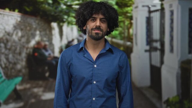 Man pointing finger with smiling face and visible curly hair, wearing blue shirt on a narrow sunlit street by white building and stone wall; joy casual friendliness.