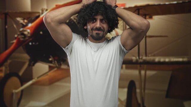 Man with curly hair and beard, hands on head beside vintage biplane in museum exhibit, wearing white tshirt and showing biceps; nostalgia amusement.