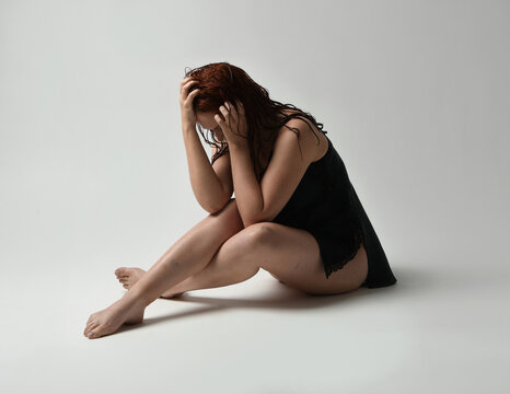 full length portrait of beautiful female model, wet messy hair, simple black silk robe gown, bruised legs. sitting pose with emotive sad pose. isolated figure studio background, dark contrast shadows