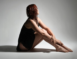 Obraz premium full length portrait of beautiful female model, wet messy hair, simple black silk robe gown, bruised legs. sitting pose with emotive sad pose. isolated figure studio background, dark contrast shadows