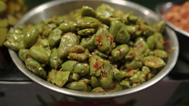 Closeup Bowl Of Fava Beans At Street Market, Glossy Pickled Beans Tossed With Chili Flakes In Metal Tray, Warm Ambient Light, Blurred Neighboring Stalls, Appetizing Green Texture, Urban