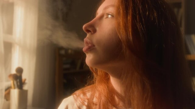 Cinematic side view shot of red haired young woman vaping and blowing smoke at home with soft yellow sunlight illuminating face, copy space