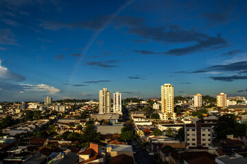 Urban landscape in Brazil during golden hour. A faint rainbow arcs across a deep blue sky above tall residential apartment buildings and typical suburban neighborhood houses