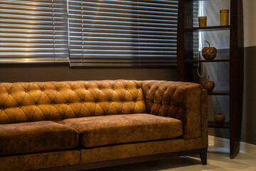 An elegant brown suede Chesterfield sofa sits in a modern office or living room. Light filters through horizontal blinds, creating a calm, sophisticated atmosphere. 