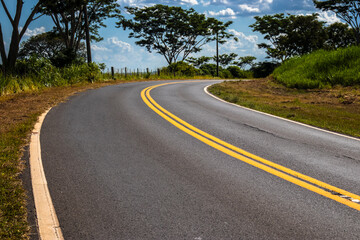 A winding asphalt road with double yellow lines curves through a lush Brazilian countryside under a partly cloudy sky, representing a journey, path, and the road ahead.