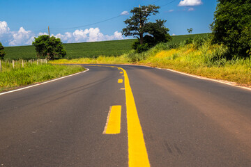 Asphalt road with yellow lines curves through the vibrant green landscape of rural Brazil under a blue sky. A concept of journey, travel, and perspective in South America's countryside.