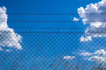 A chain-link fence with barbed wire under a blue sky with clouds. It symbolizes security, borders, division, danger, and the contrast between confinement and freedom.