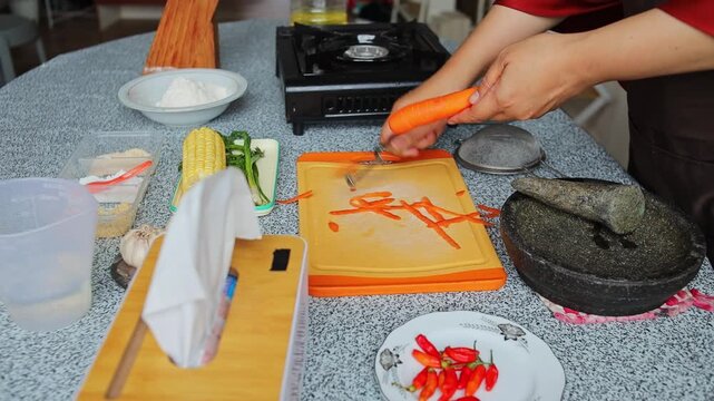 Close up of hand home chef peeling fresh carrot with handheld peeler on orange cutting board in home kitchen
