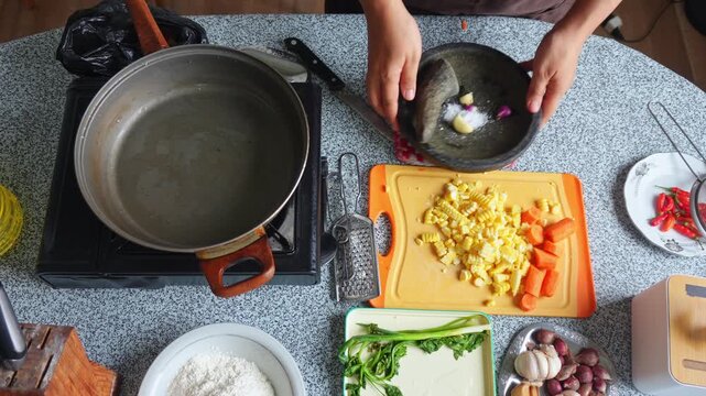 Overhead shot of hands grinding garlic and shallots in traditional stone mortar for Indonesian corn fritters