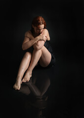 Naklejka premium full length female model with damp hair, wearing simple black glamour dress. sitting kneeling pose on wet black floor with water reflections. isolated figure on dark studio background.