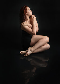 full length female model with damp hair, wearing simple black glamour dress. sitting kneeling pose on wet black floor with water reflections. isolated figure on dark studio background.