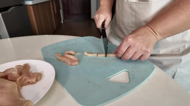 The housewife prepares dinner, cuts chicken fillet. Women's hands with a knife and a cutting board on a home kitchen table