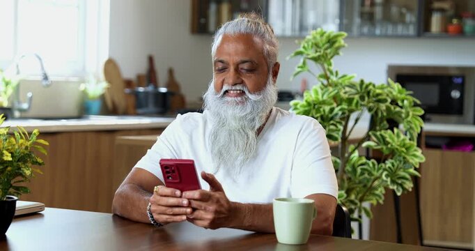 Indian mid age stylish man with long white beard and hair using smartphone while sitting at breakfast table in modern kitchen interior, browsing internet, chatting online at home