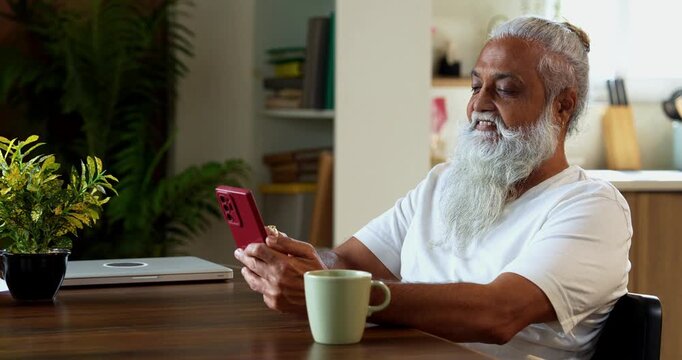 Indian mid age stylish man with long white beard and hair using smartphone while sitting at breakfast table in modern kitchen interior, browsing internet, chatting online at home