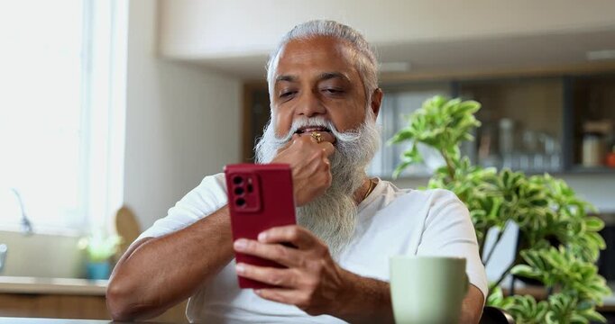 Indian mid age stylish man with long white beard and hair using smartphone while sitting at breakfast table in modern kitchen interior, browsing internet, chatting online at home