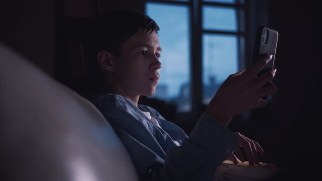 Peaceful Evening: Boy Enjoying Solitude by the Window, Snacking on Chips, and Using His Phone
