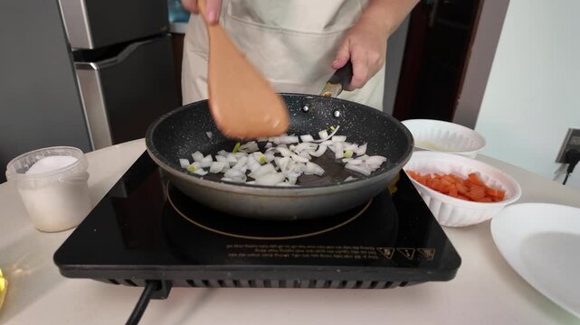 Woman is frying onions and carrots in an induction frying pan, stirring with a wooden spatula