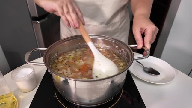 A woman cooks soup in a pot on an induction hob, stirring the soup with a ladle