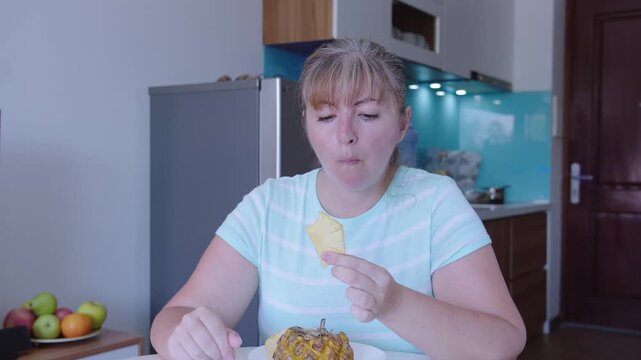 A woman is sitting at a table in the background of the kitchen and eating pineapple