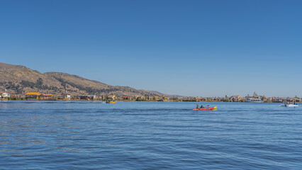 Fototapeta premium Tourist boats, reed boats balsa de totora sail on a calm alpine lake. Huts are visible on the shore of Uros Floating Island. Mountains against a clear blue sky. Peru. Puno. Lake Titicaca. Copy space