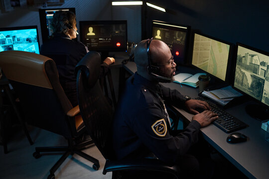 Black man wearing headset monitoring security footage on multiple computer screens in control room, Caucasian middle aged woman working at adjacent station in background