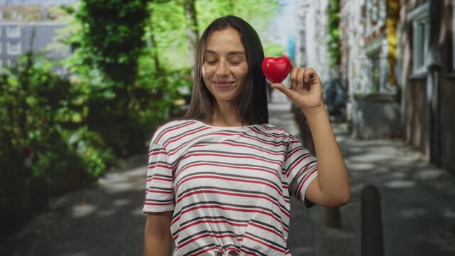 Young hispanic woman holds red heart in hand on street beside face wearing striped shirt with knotted hem and raised arm, eyes softly closed; love joy.