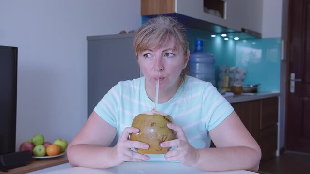 Woman drinks juice from a fresh coconut through a straw, sits at a table