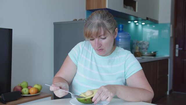 A woman eats avocado pulp with a spoon, sits at a table