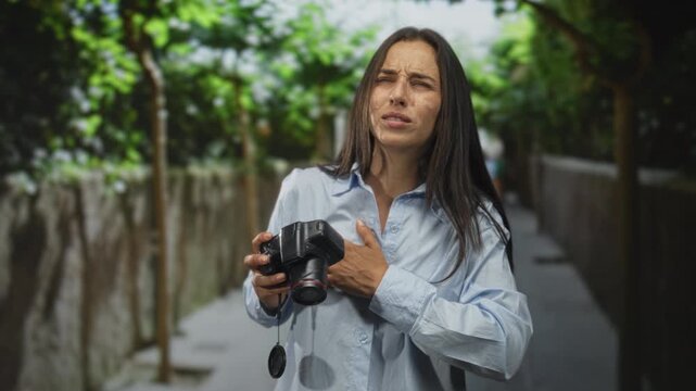 Woman holding camera clutching chest on street, grimacing with closed eyes and lens cap dangling, appearing to have heartattack; pain.